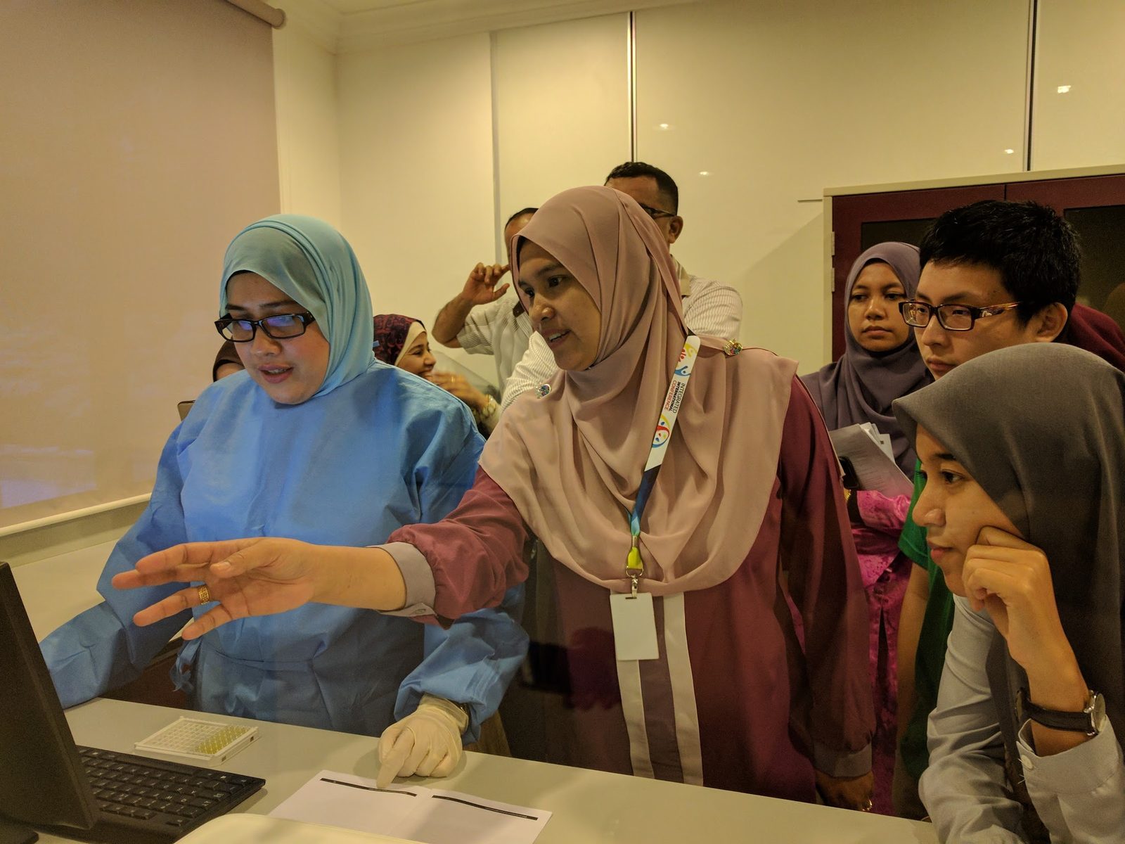 A trainer in a blue lab gown points at a laptop screen next to a 96-well plate as participants look on