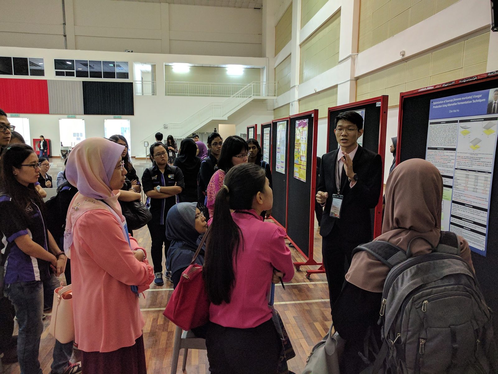 A presenter in a dark suit explains his research poster to a group of visitors in the hall