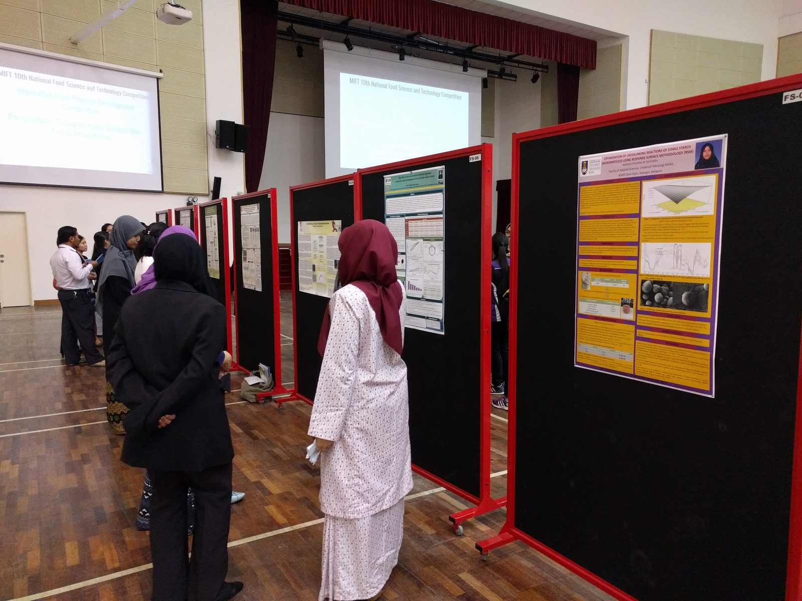 Visitors reading research posters on red-framed boards in a large university hall