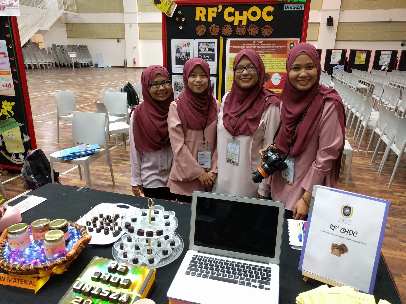 Four students in matching outfits stand behind their chocolate product booth, with a laptop and samples