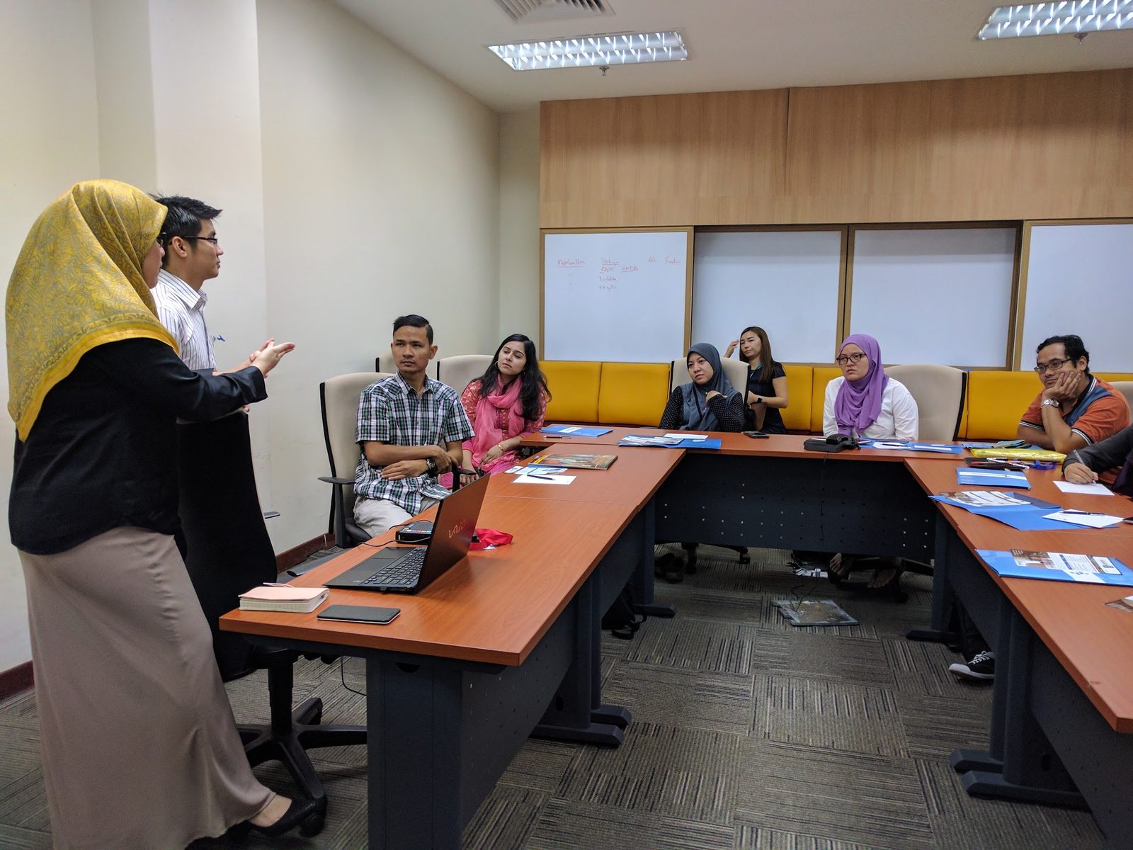 Two speakers stand at the front of a meeting room while participants at a U-shaped table listen