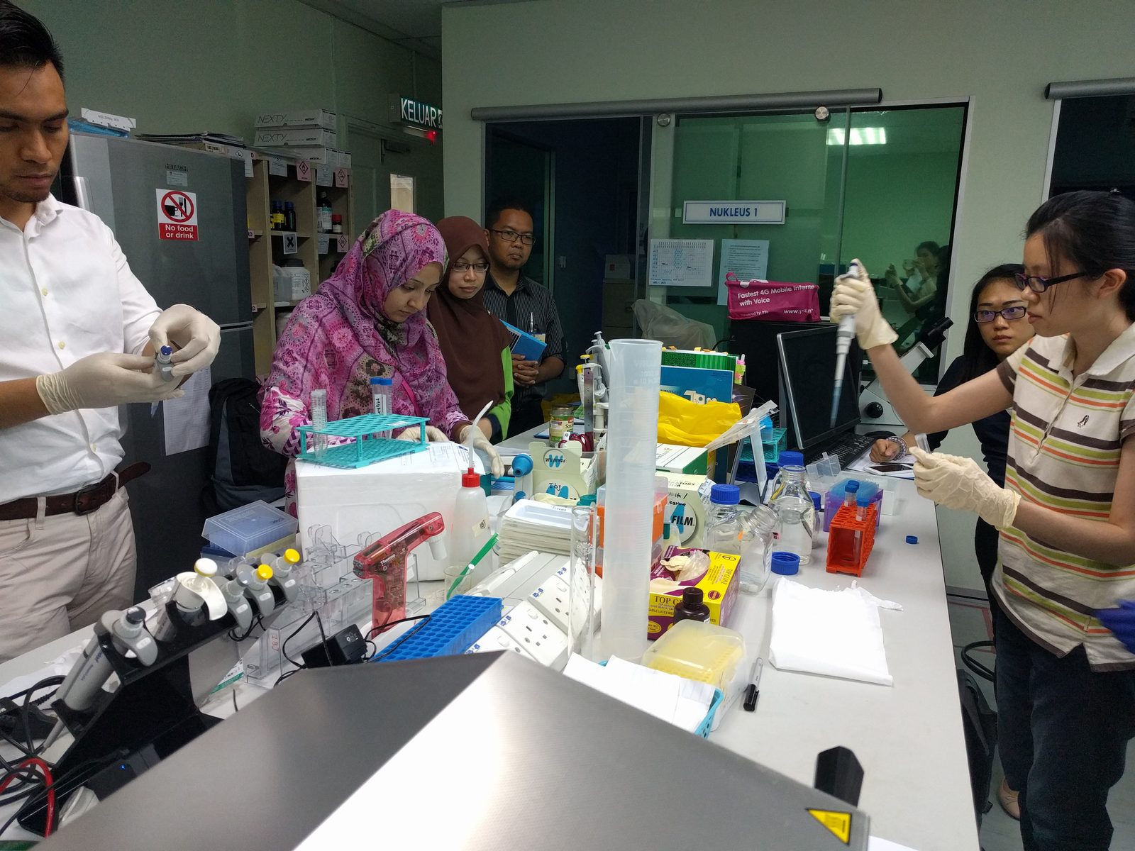 Gloved hands pipette samples into tubes at a busy laboratory bench while others watch
