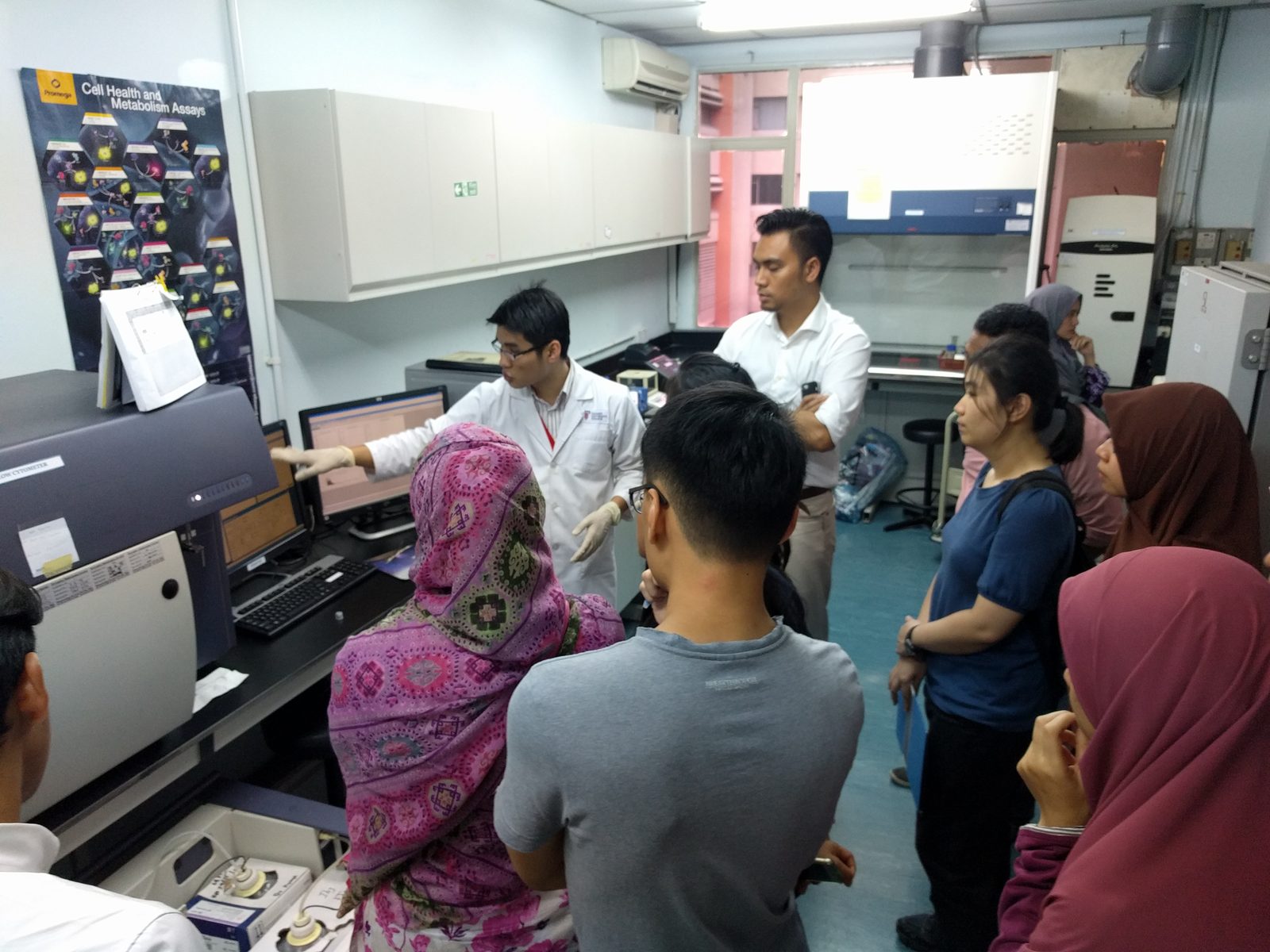 A presenter in a lab coat points to the flow cytometer screen while participants watch