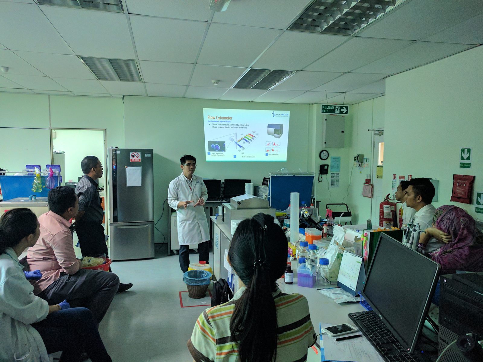A presenter in a lab coat gives a talk beside a projected slide titled Flow Cytometer