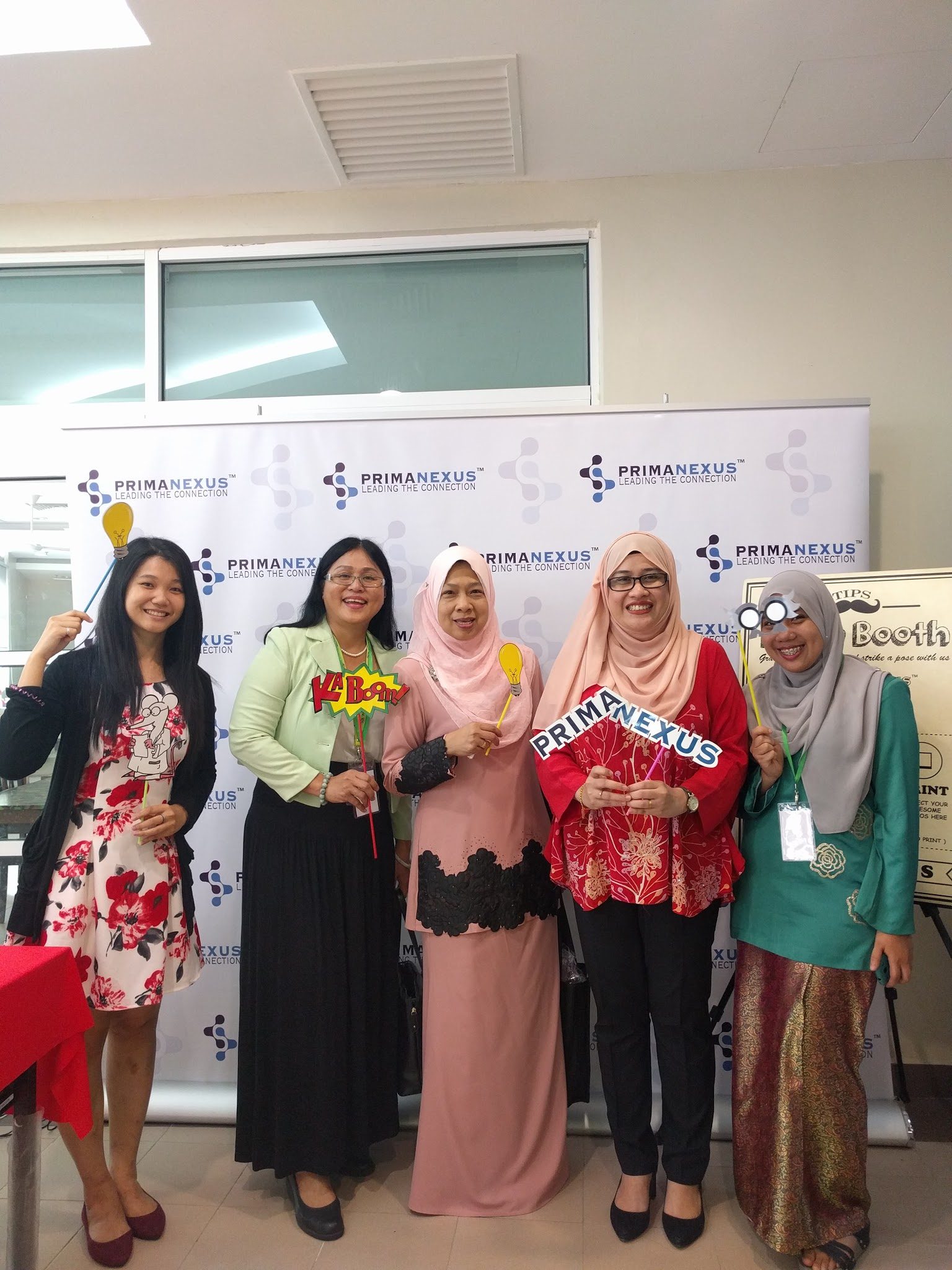 Five women hold photo props in front of the Prima Nexus backdrop at the photo booth