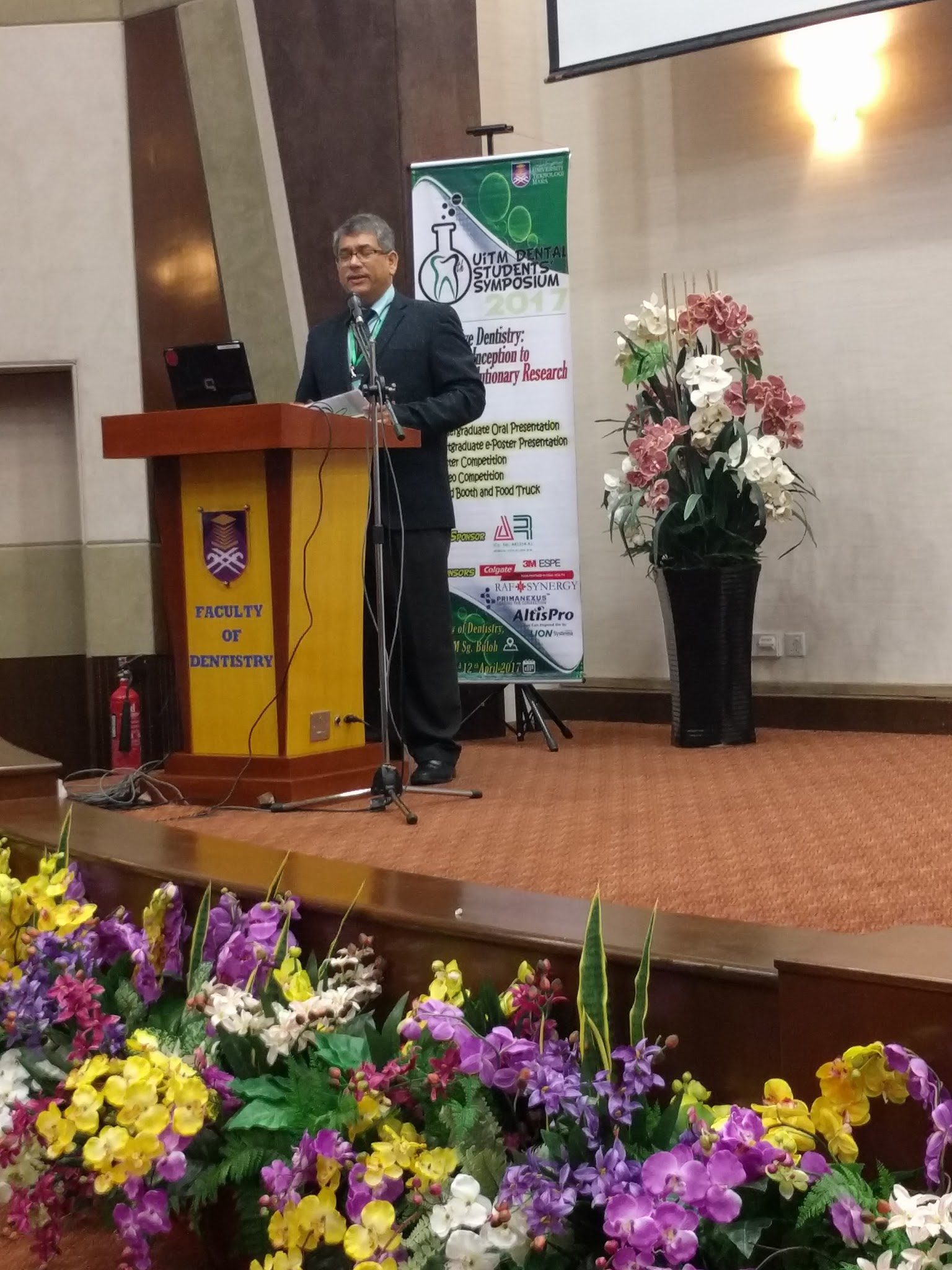 A speaker at the lectern on the symposium stage, beside the event banner and a flower arrangement