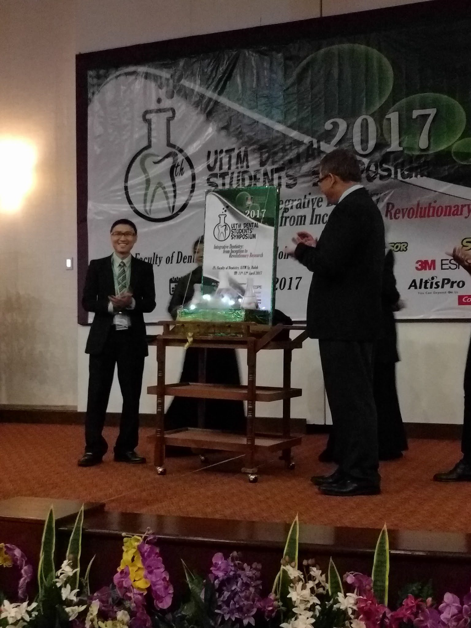 Two men in suits on stage beside a glass display on a trolley, in front of the symposium backdrop