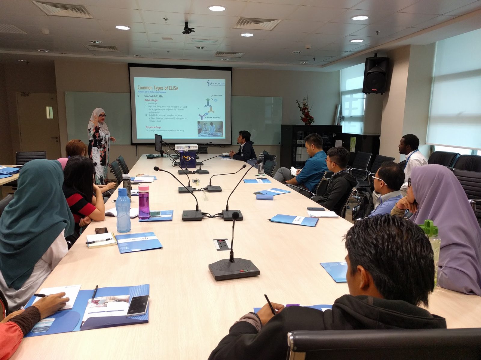 A presenter explains a slide on common types of ELISA to participants seated around a meeting table