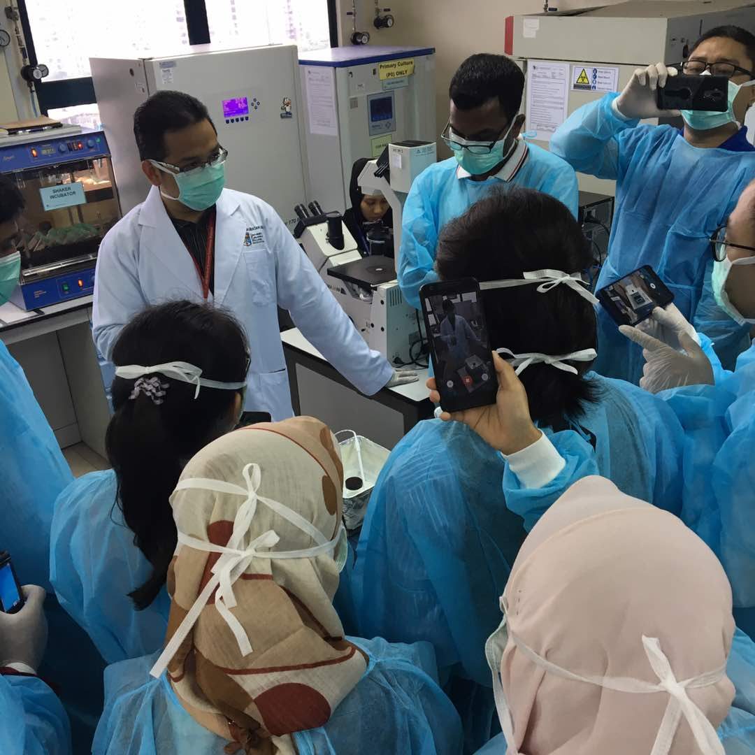 A demonstrator in a white coat at a lab bench while participants in gowns record on their phones