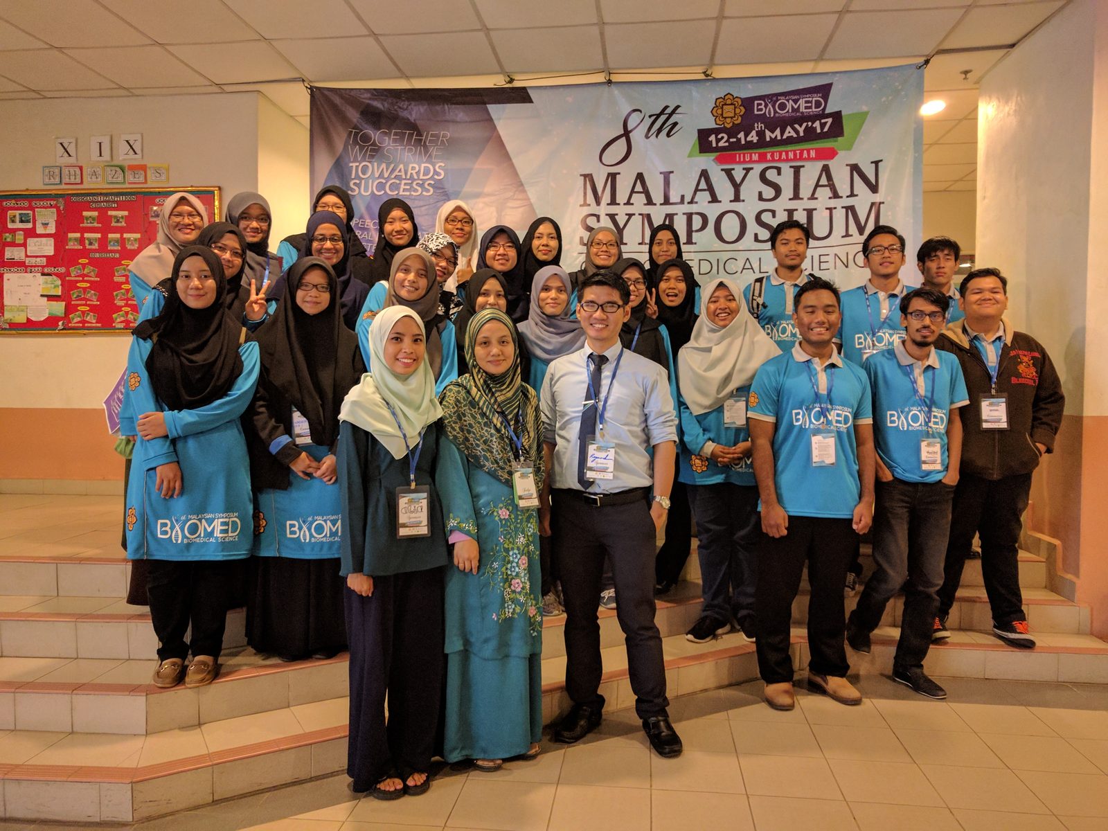 A large group, many in blue symposium T-shirts, poses in front of the 8th Malaysian Symposium banner.