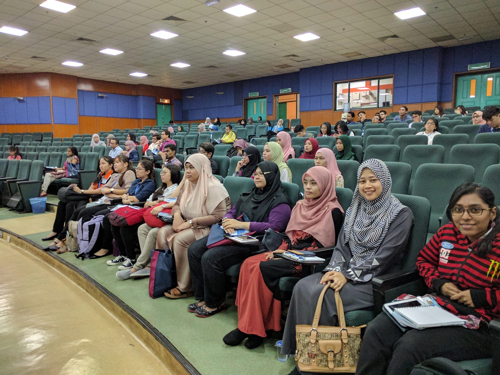 Participants seated in rows in a lecture hall, some with notes on their laps.