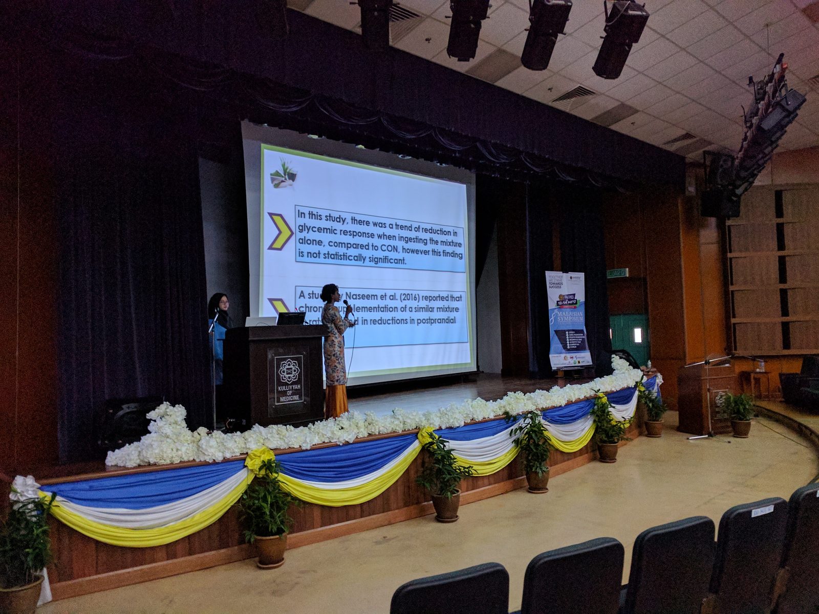 A presenter at the lectern on the hall stage, with slides on the screen behind.