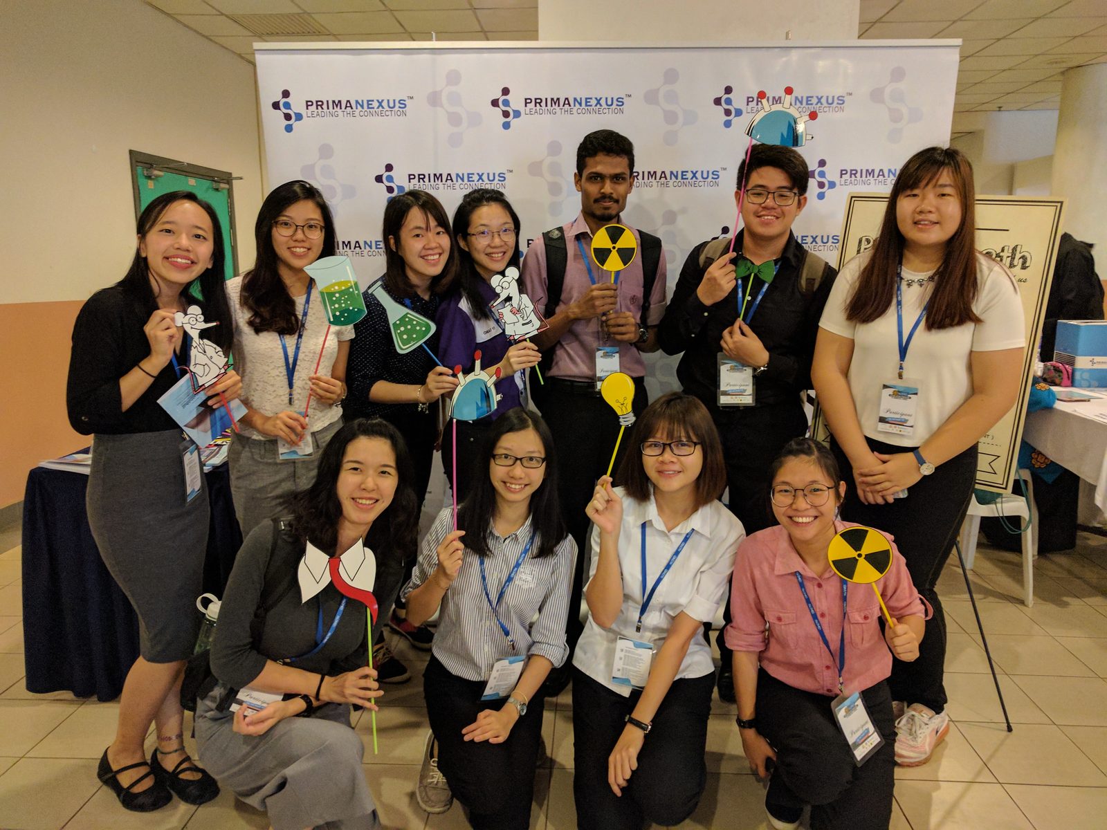 Smiling participants hold science-themed photo props in front of a Prima Nexus backdrop.