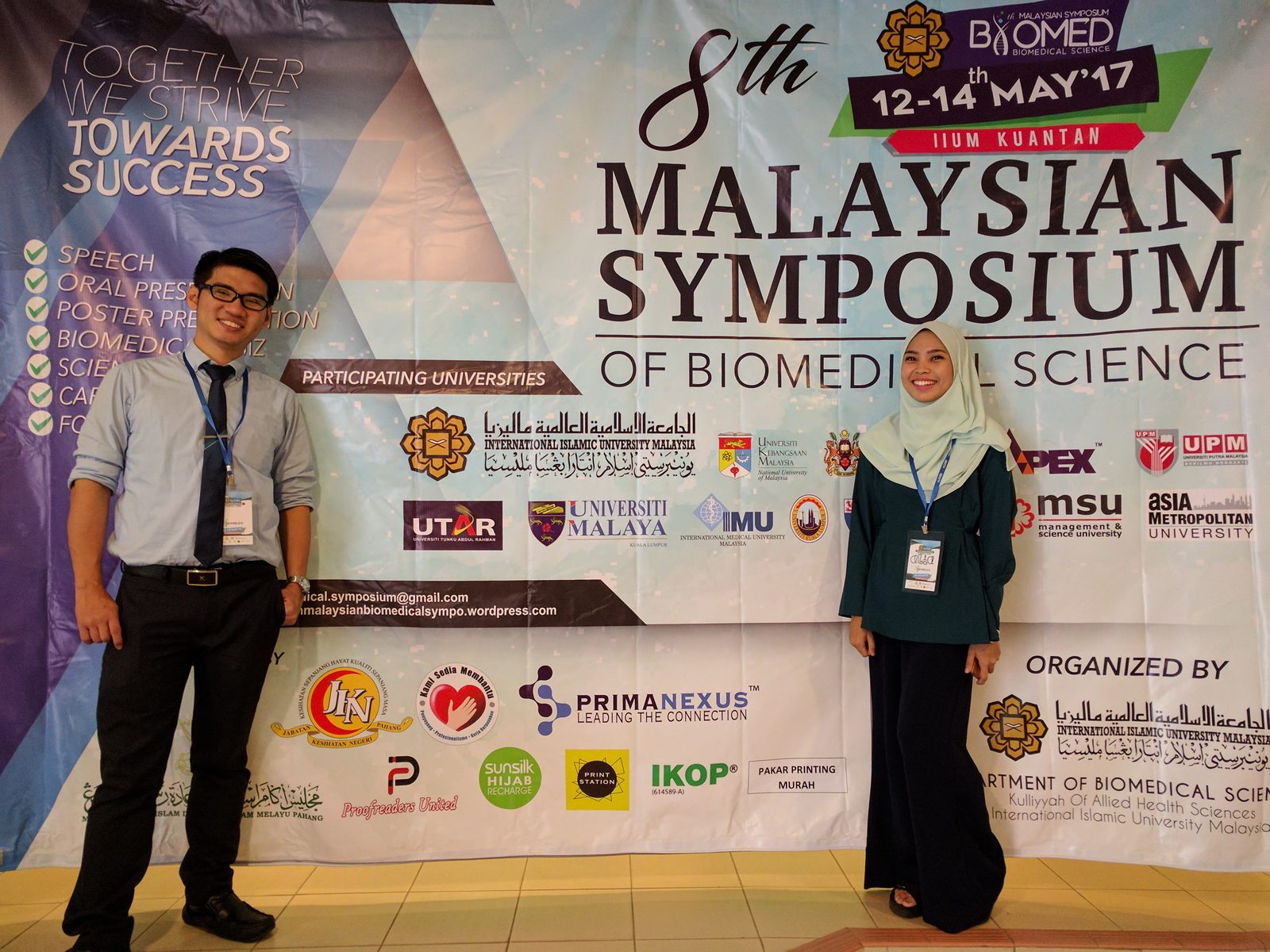 Two people stand beside the symposium banner, which carries the Prima Nexus logo.