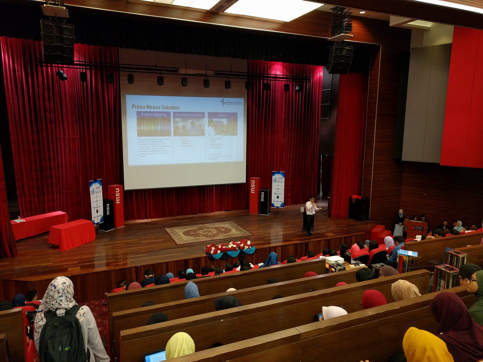 Speaker on the theatre stage with a Prima Nexus slide on the screen and students seated in rows