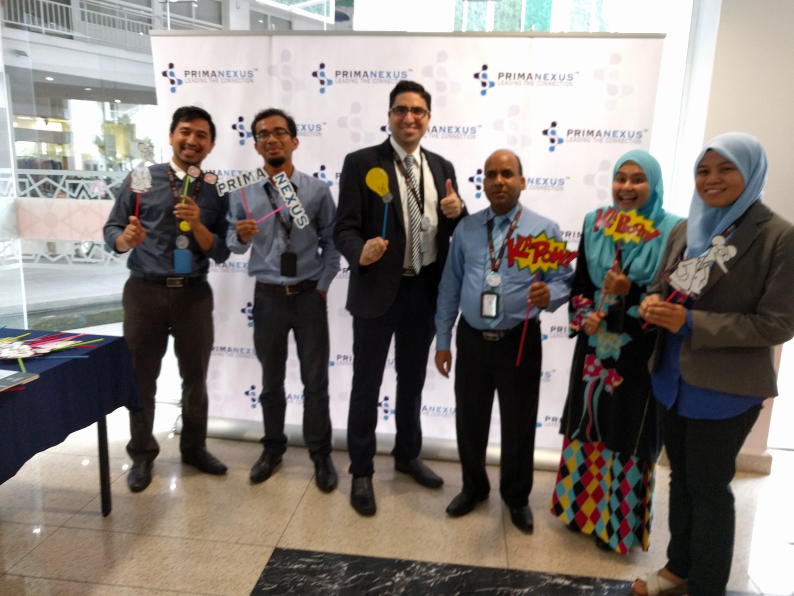 Six participants holding photo props in front of the Prima Nexus backdrop