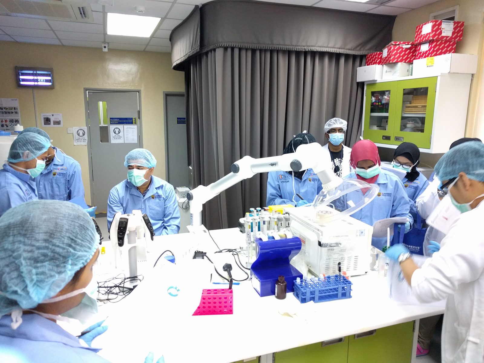 Participants in lab coats, masks and hair covers gather around a laboratory bench with pipettes and sample racks