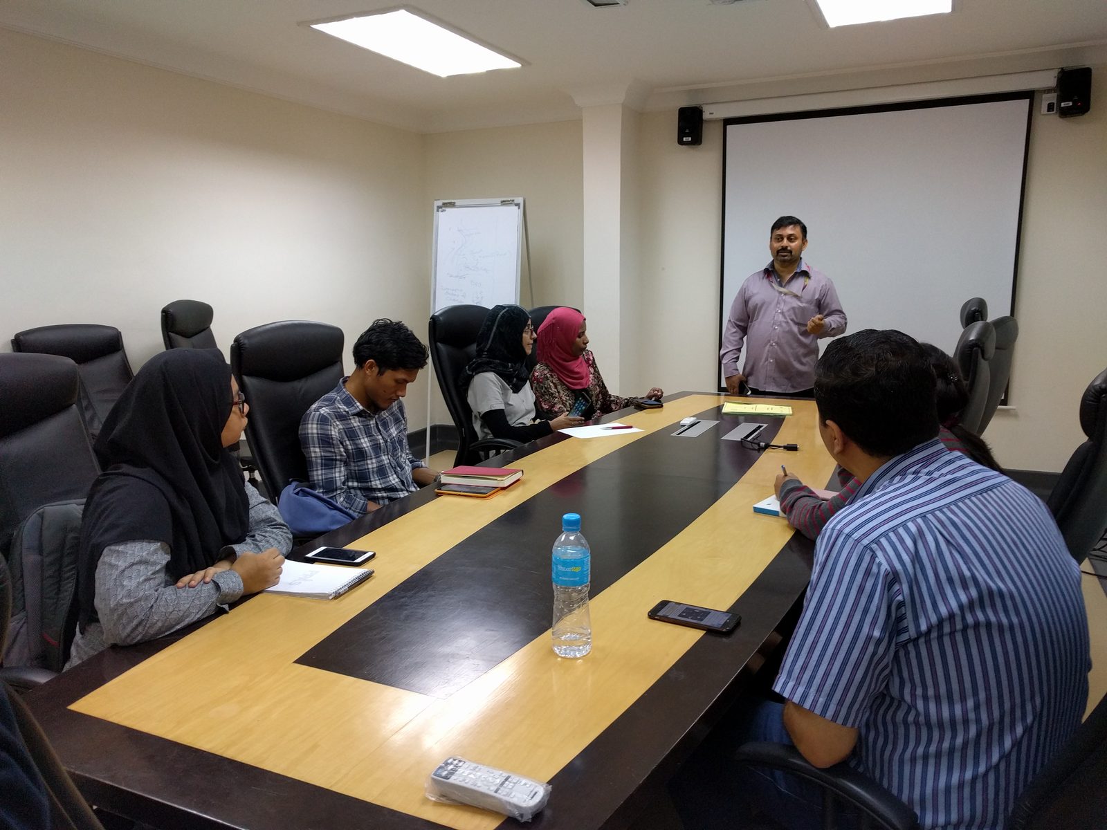 A speaker stands at the front of a meeting room while participants sit around the table