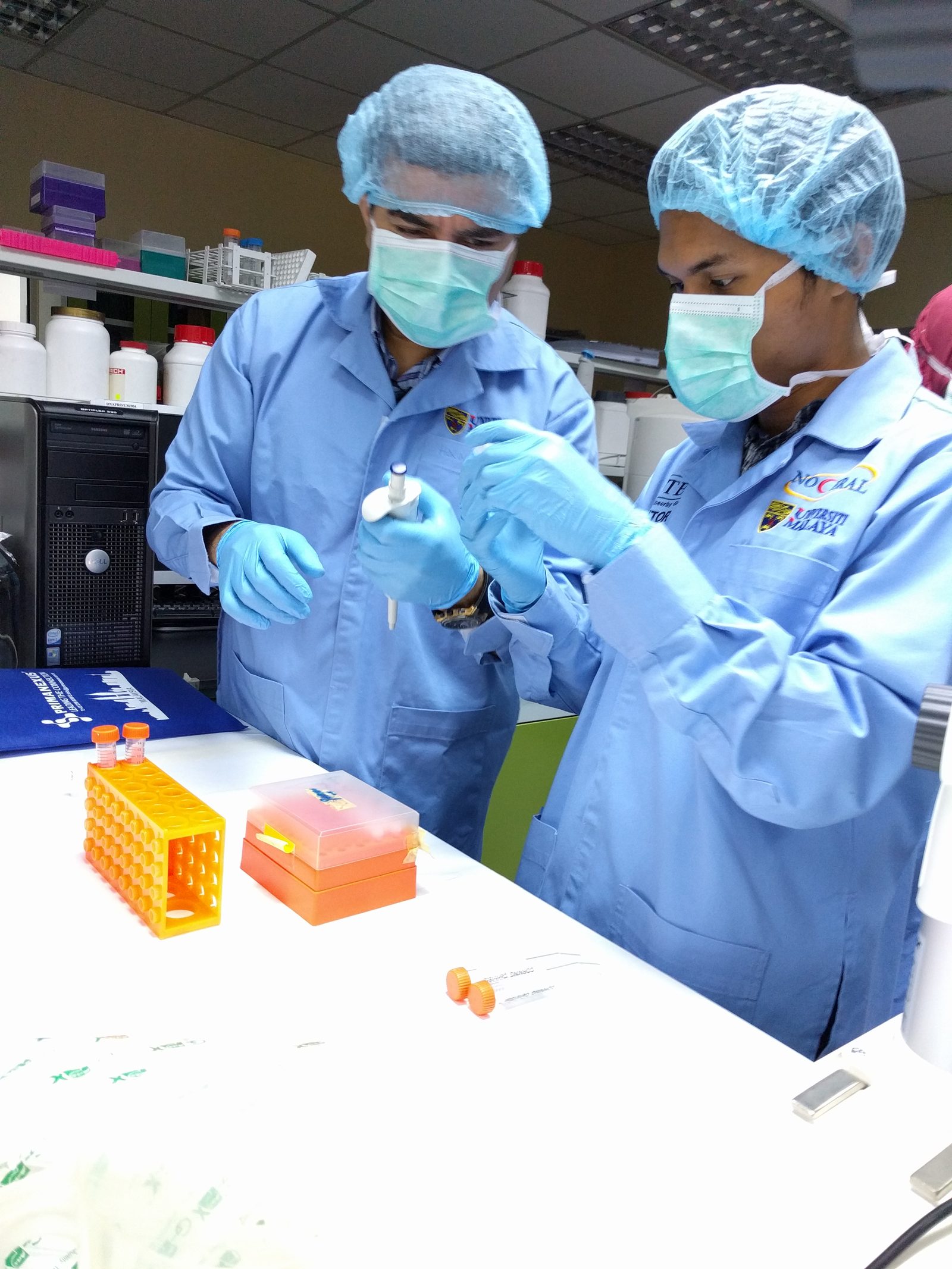 Two participants in hair covers and masks handle a pipette and sample tubes at the bench