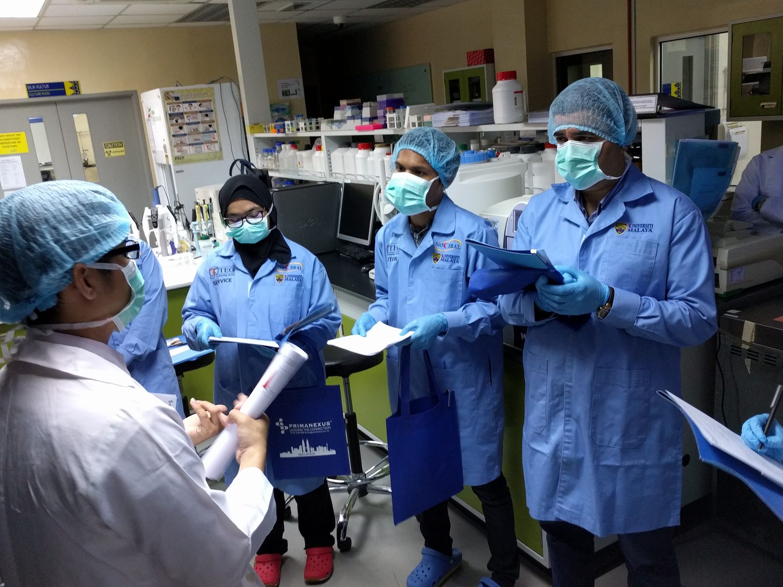 Participants in blue lab coats hold folders and papers during a briefing in the laboratory