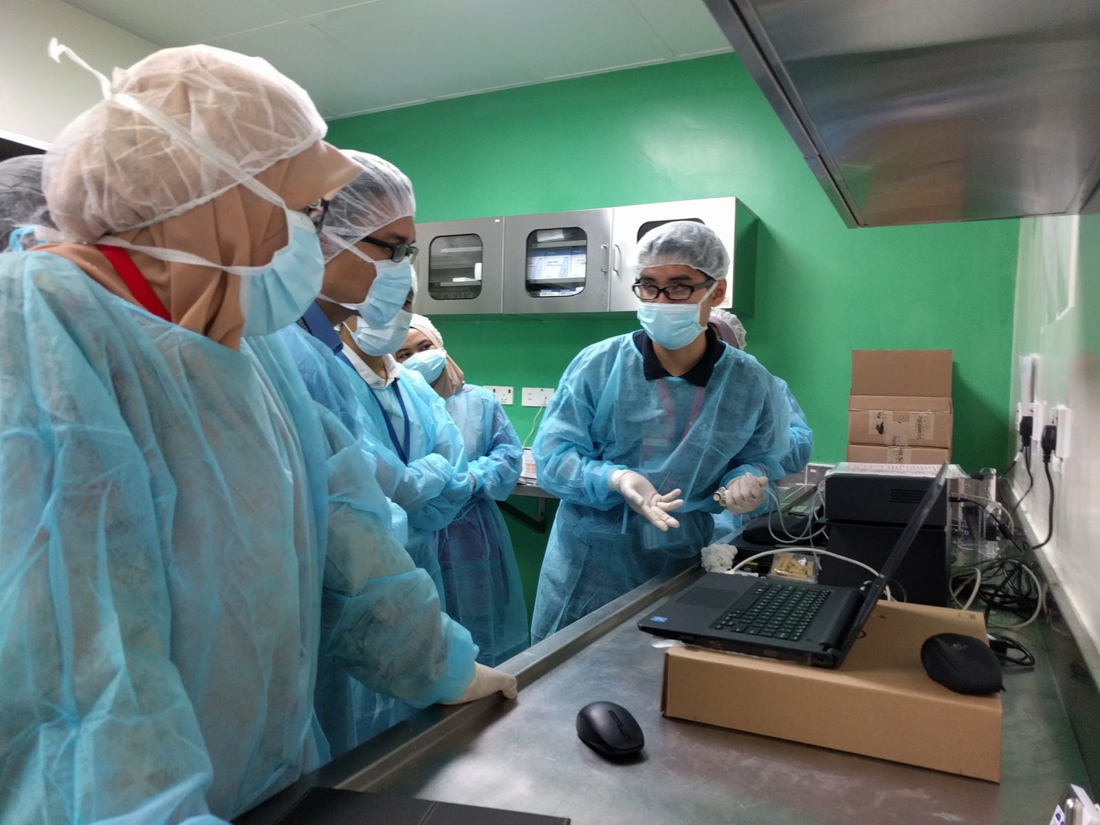 A trainer in gown and mask demonstrates the CODA system at a lab bench to a group of gowned participants