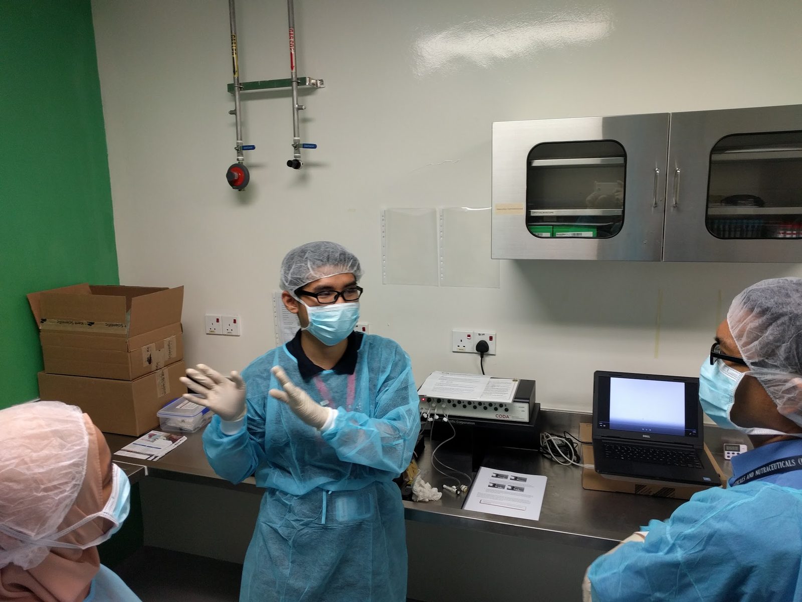 A trainer in gown and mask explains the CODA controller and laptop on a stainless steel bench