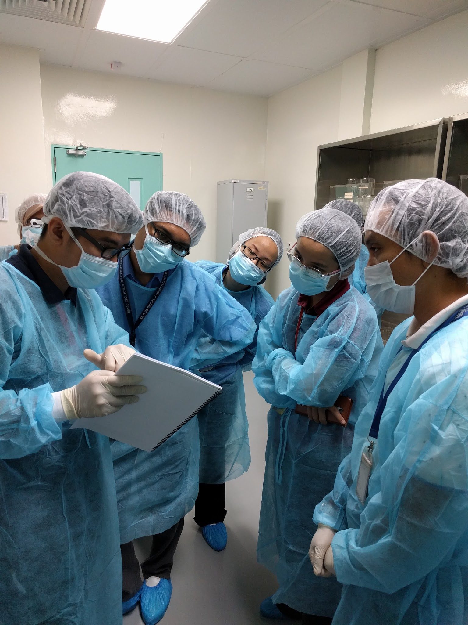 A trainer holds a spiral-bound manual while five gowned participants stand around and listen