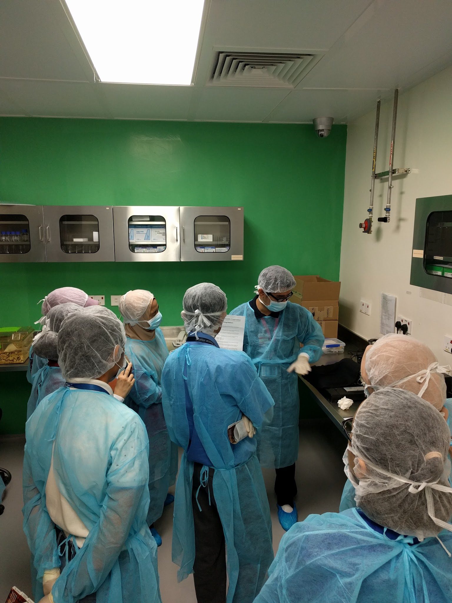 Participants in gowns and hair covers, seen from behind, gather around a trainer in the facility room