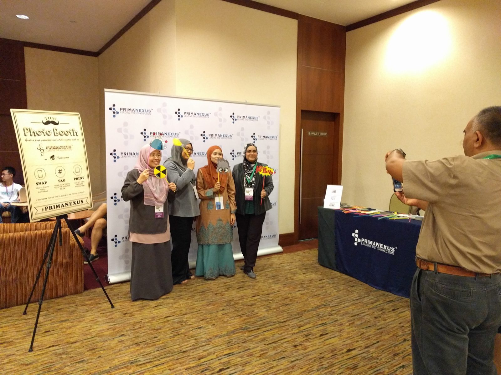 Four delegates hold photo props in front of the Prima Nexus backdrop while a man takes their picture
