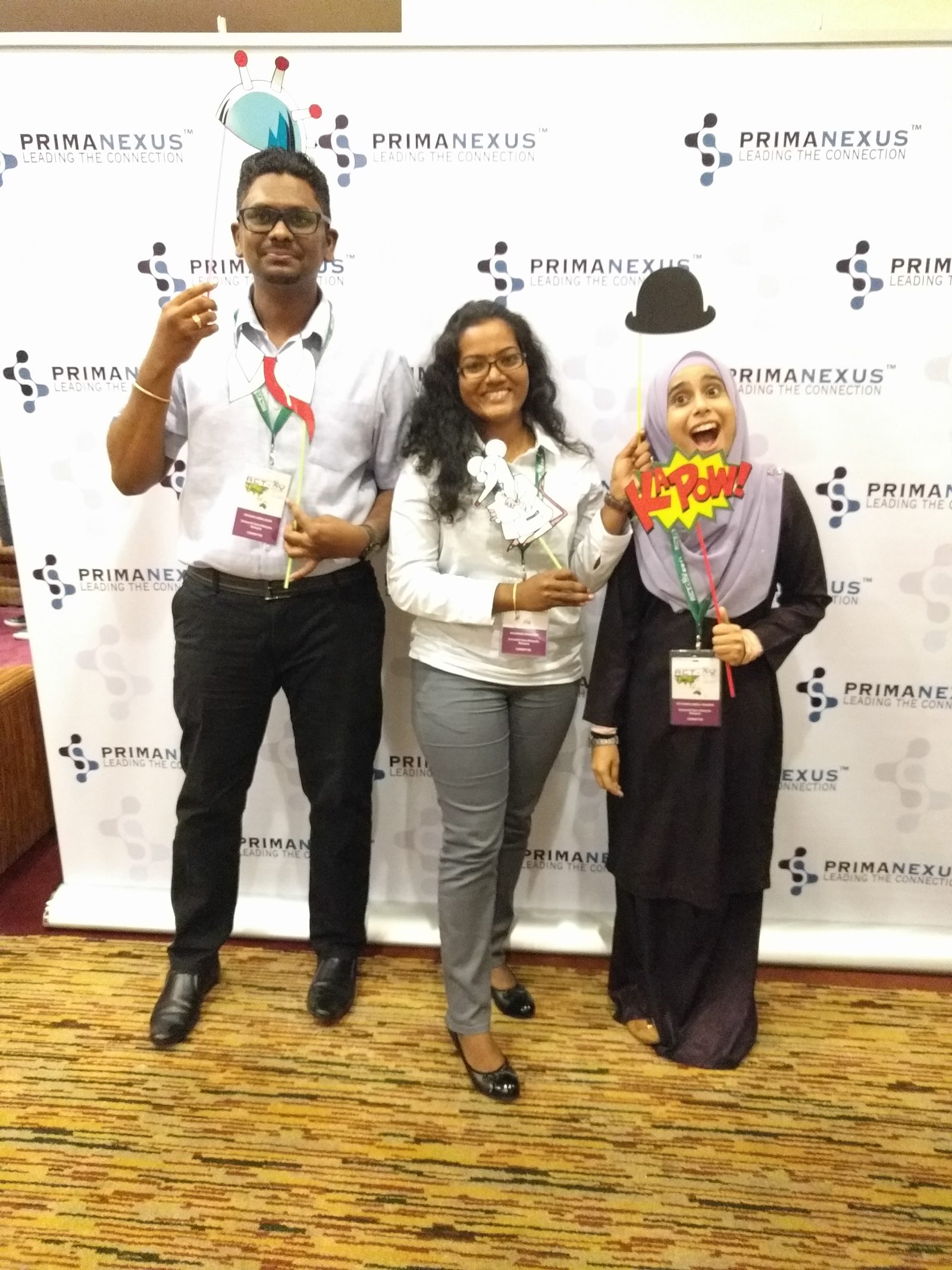 Three delegates with conference badges pose with props in front of the Prima Nexus backdrop