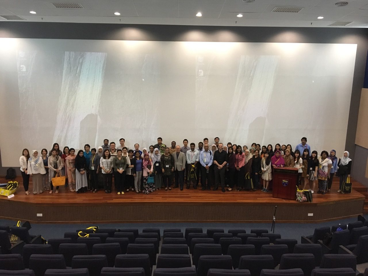 Course participants standing together on the auditorium stage for a group photo, rows of empty seats in front