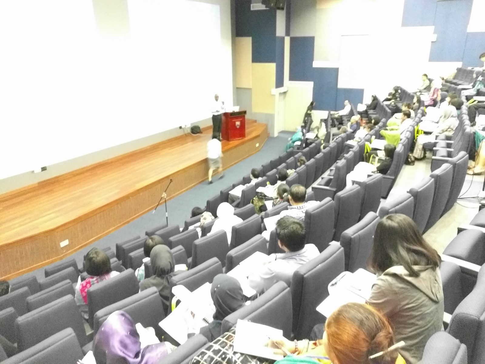 Delegates seated in the tiered auditorium while a lecturer stands at the podium on the stage
