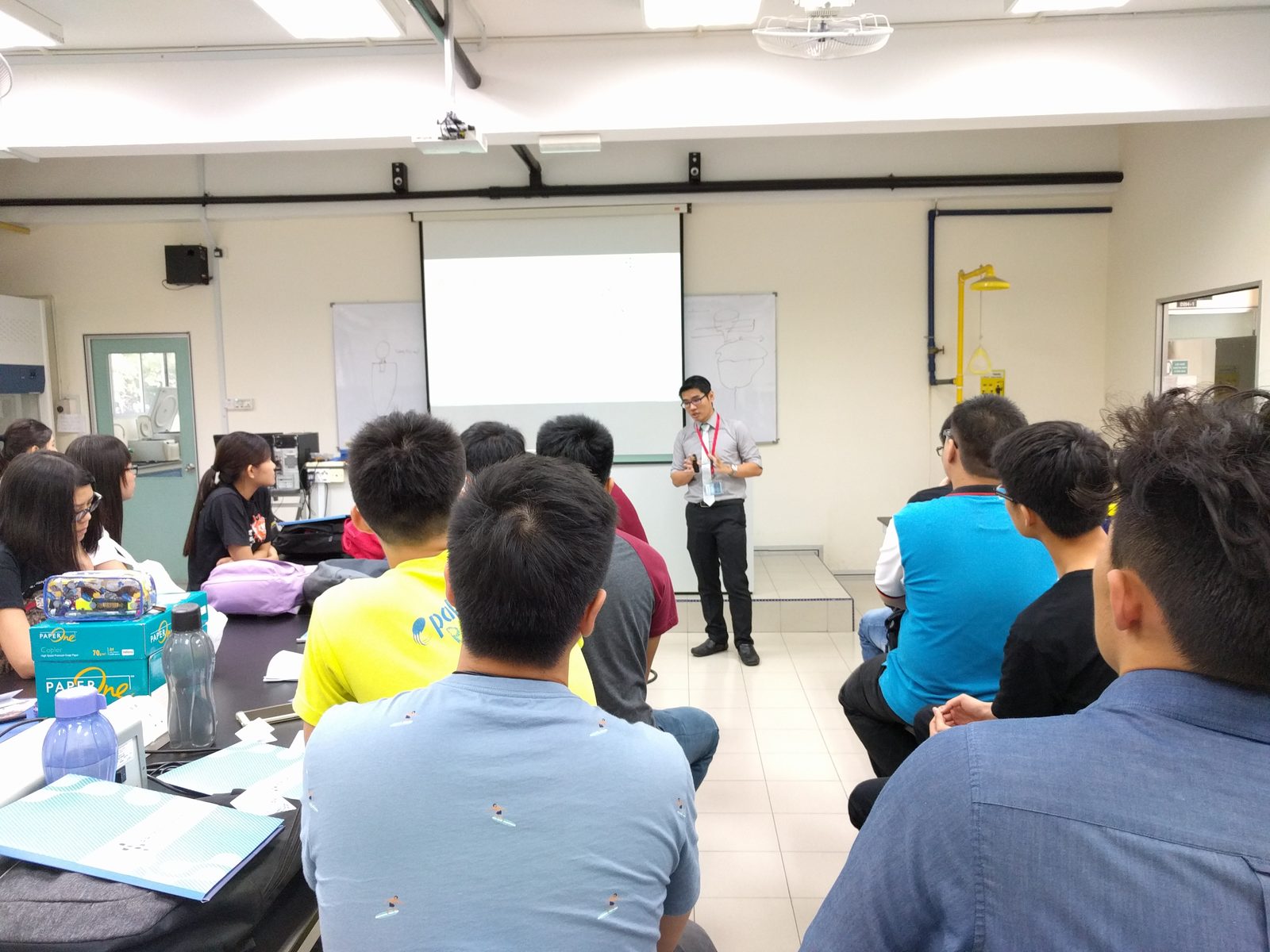 A speaker presents in front of a projection screen while participants listen from their benches