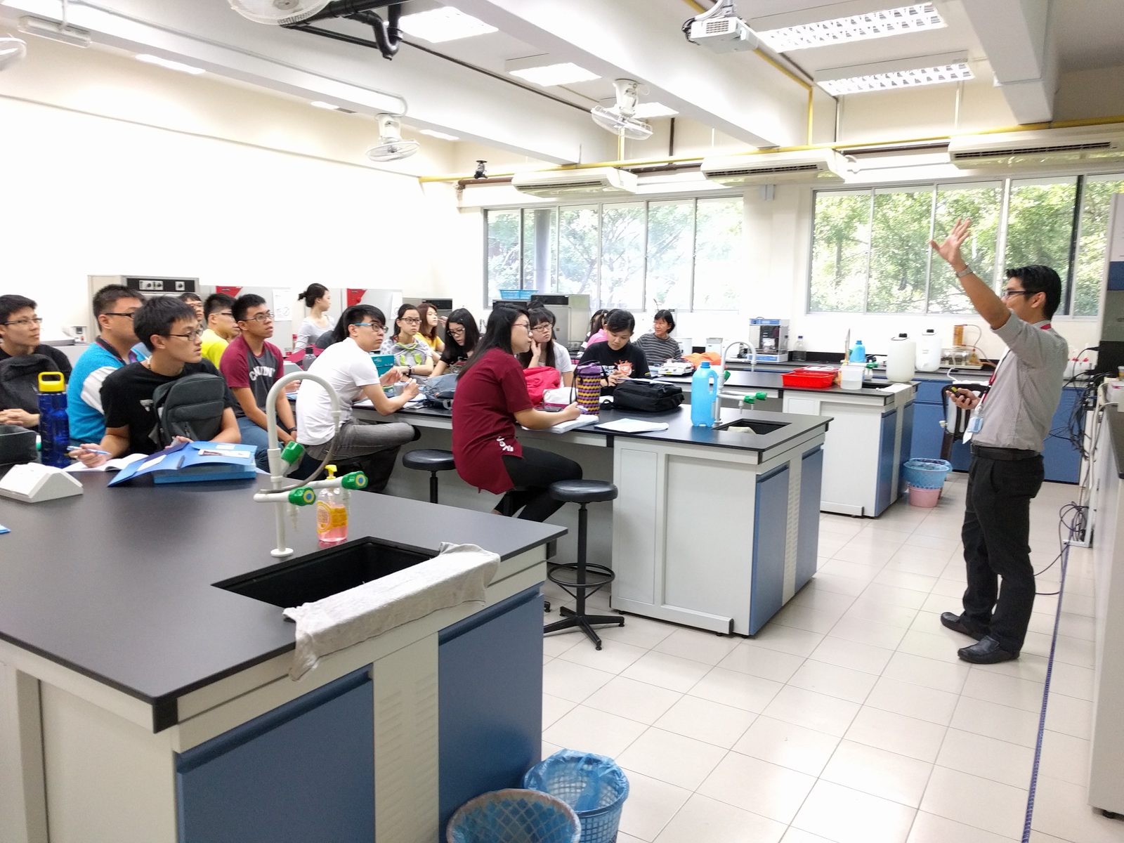 A presenter raises one hand while speaking to participants seated at laboratory benches