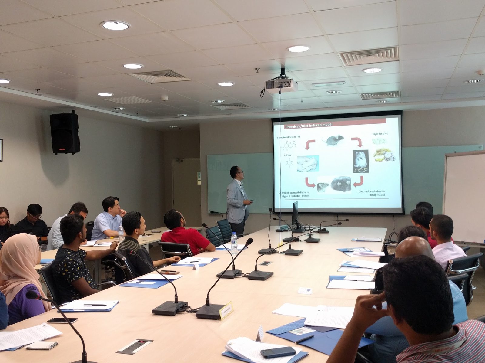 A presenter stands beside a projected slide at the front of the room, with researchers seated at long tables