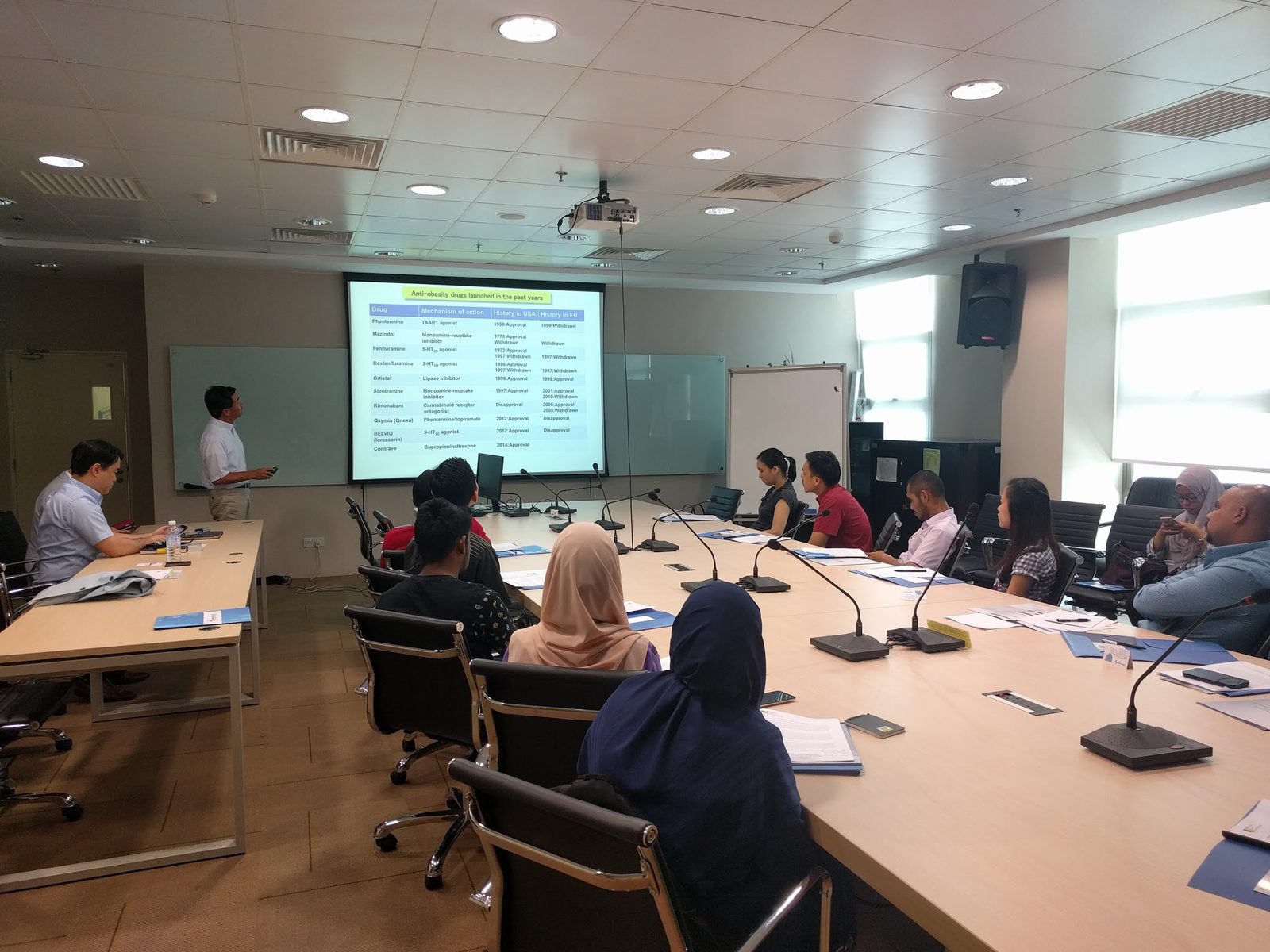 A speaker at the front of the seminar room stands beside a projected table, with participants seated at long tables