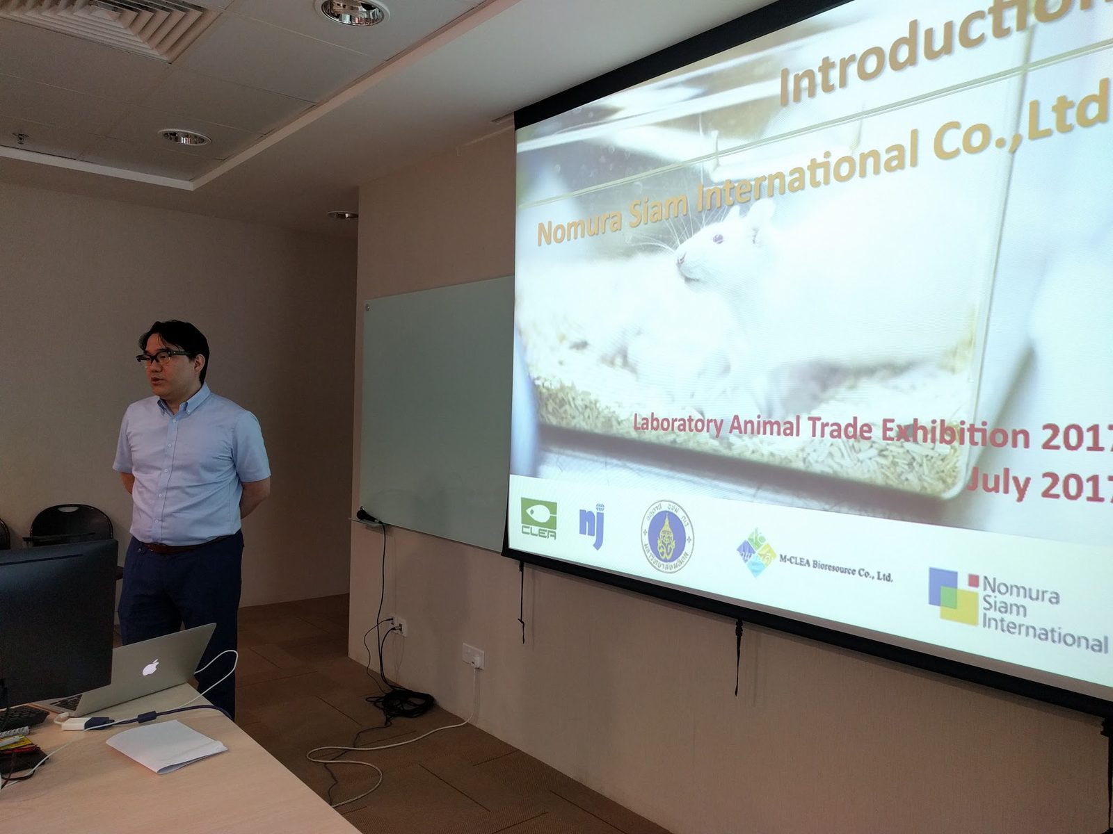 A presenter stands beside a projected slide introducing Nomura Siam International