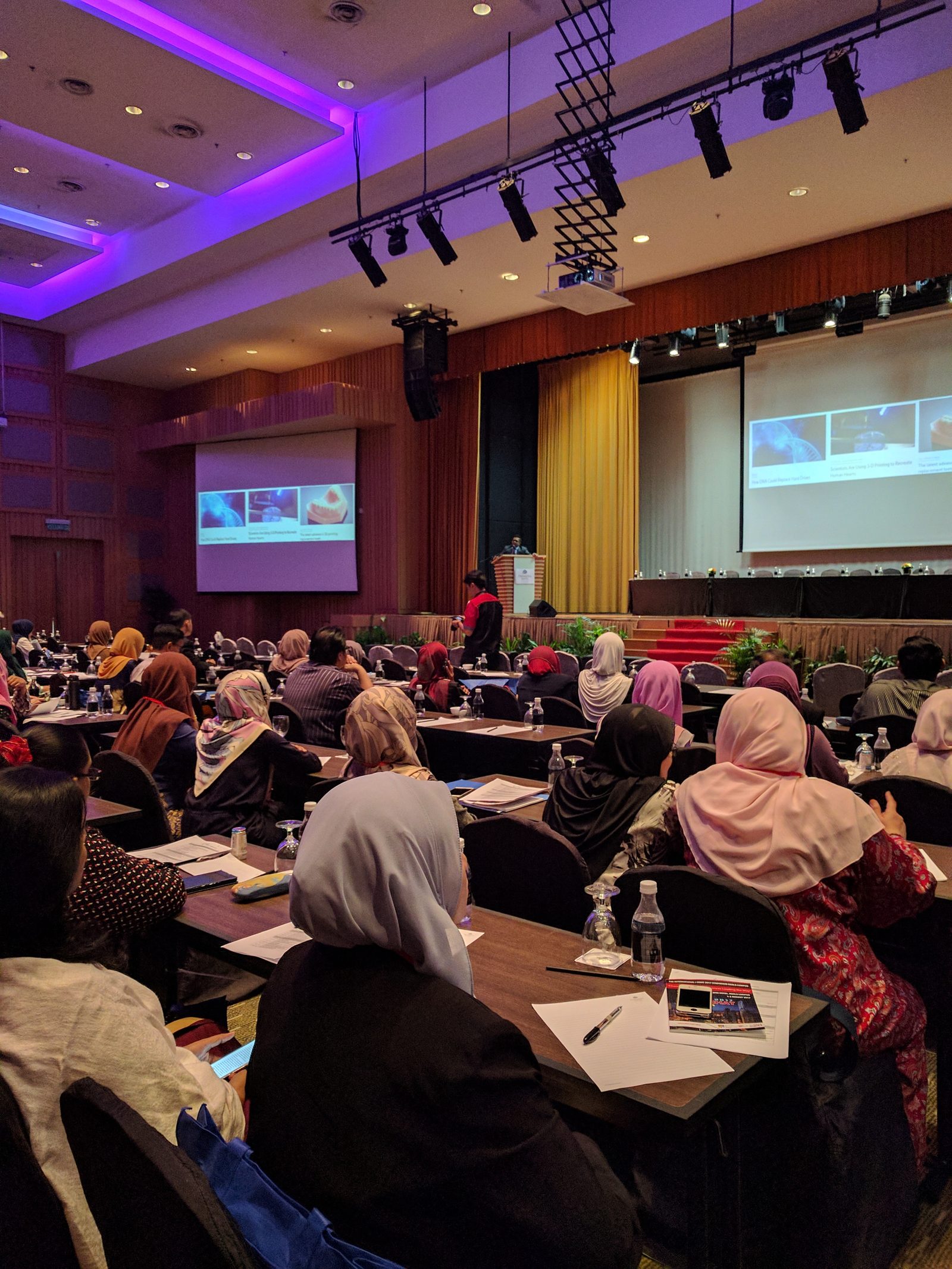 Delegates at tables listen to a talk, with slides shown on large screens beside the stage