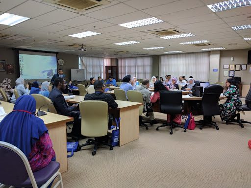 Delegates seated at desks and office chairs in a meeting room, with a projector screen at the front.