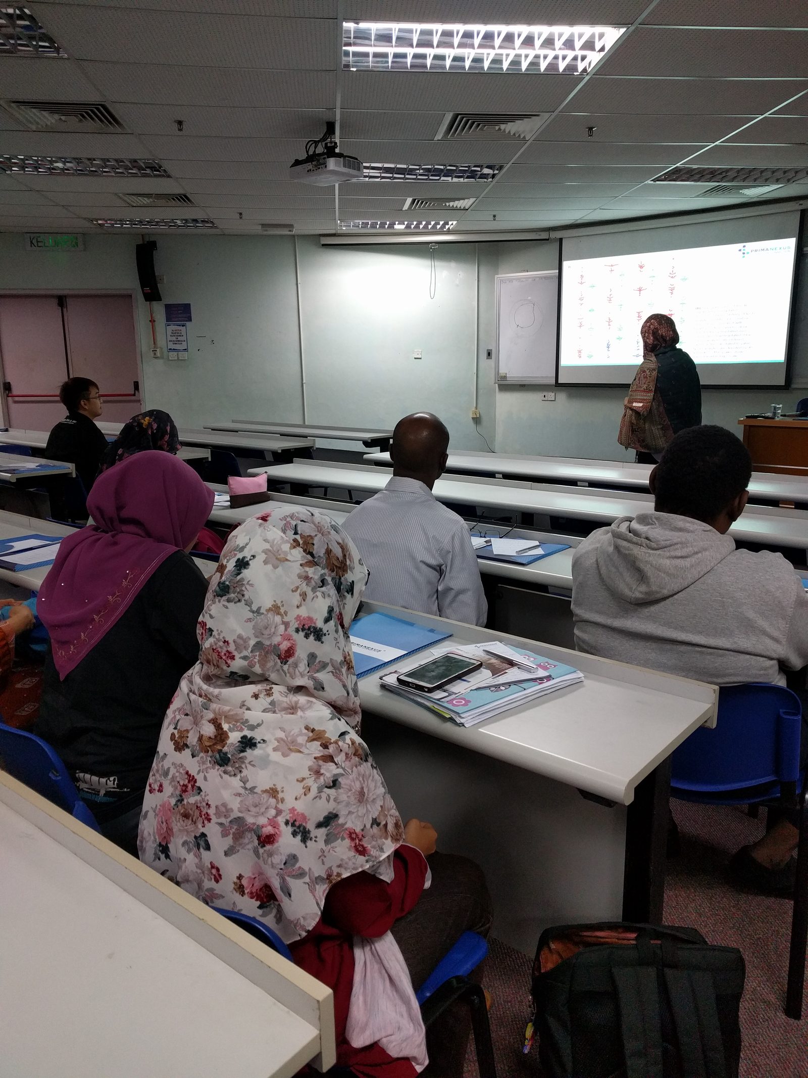 A presenter stands beside a projected slide while participants listen from their desks