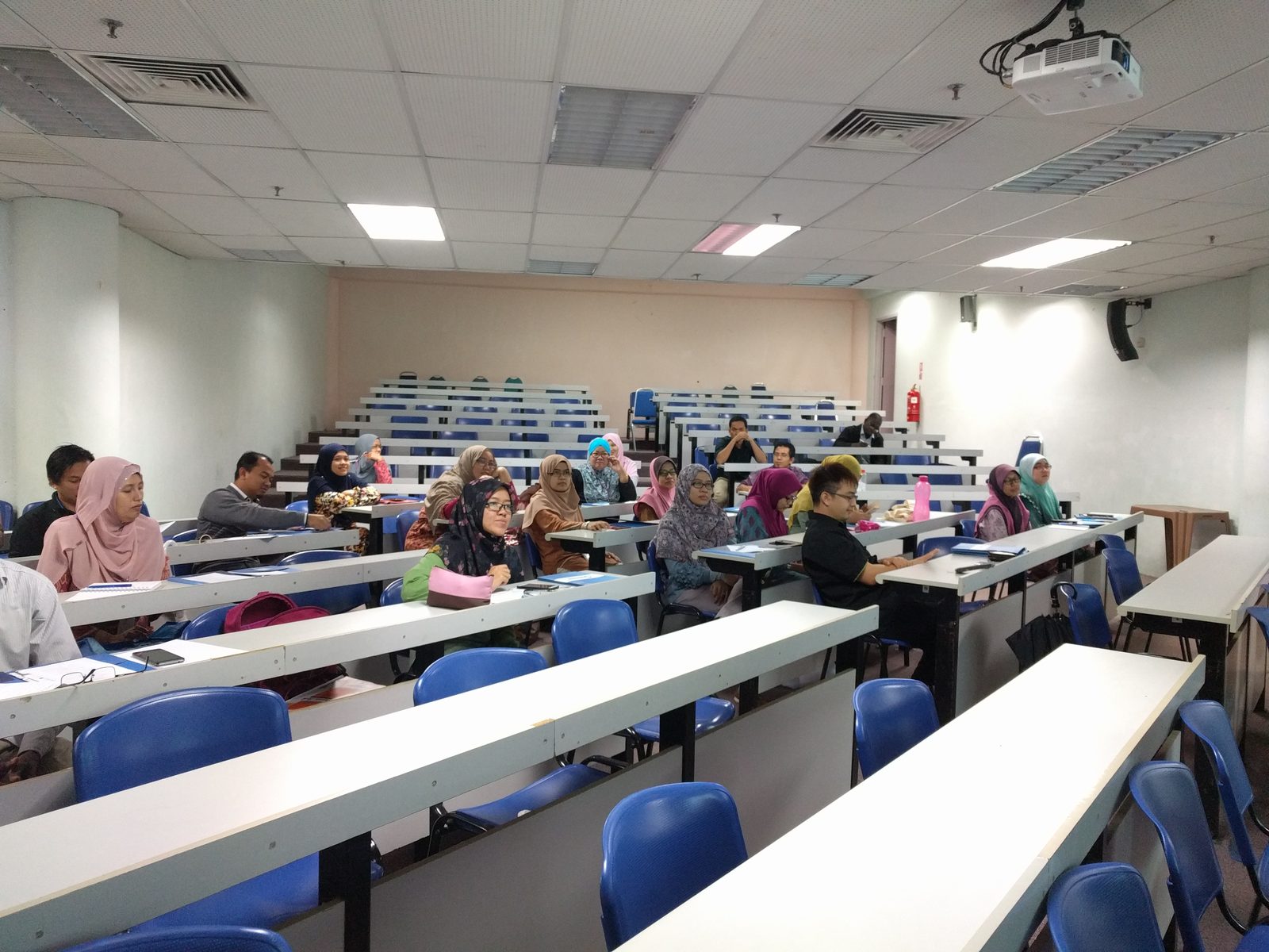 Participants seated at long desks in a tiered lecture room, facing the front