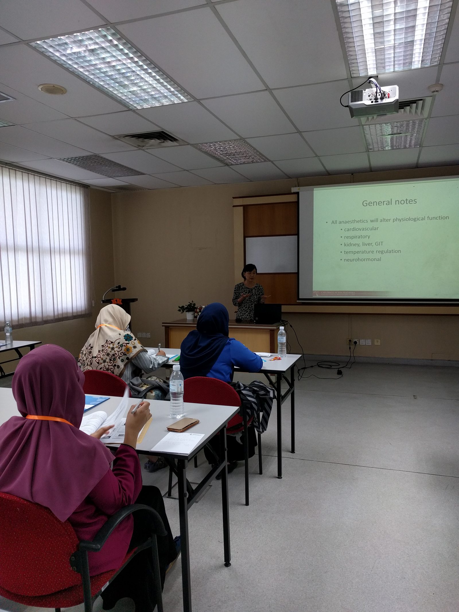 A lecture room with participants at tables facing a speaker and a slide on how anaesthetics affect the body