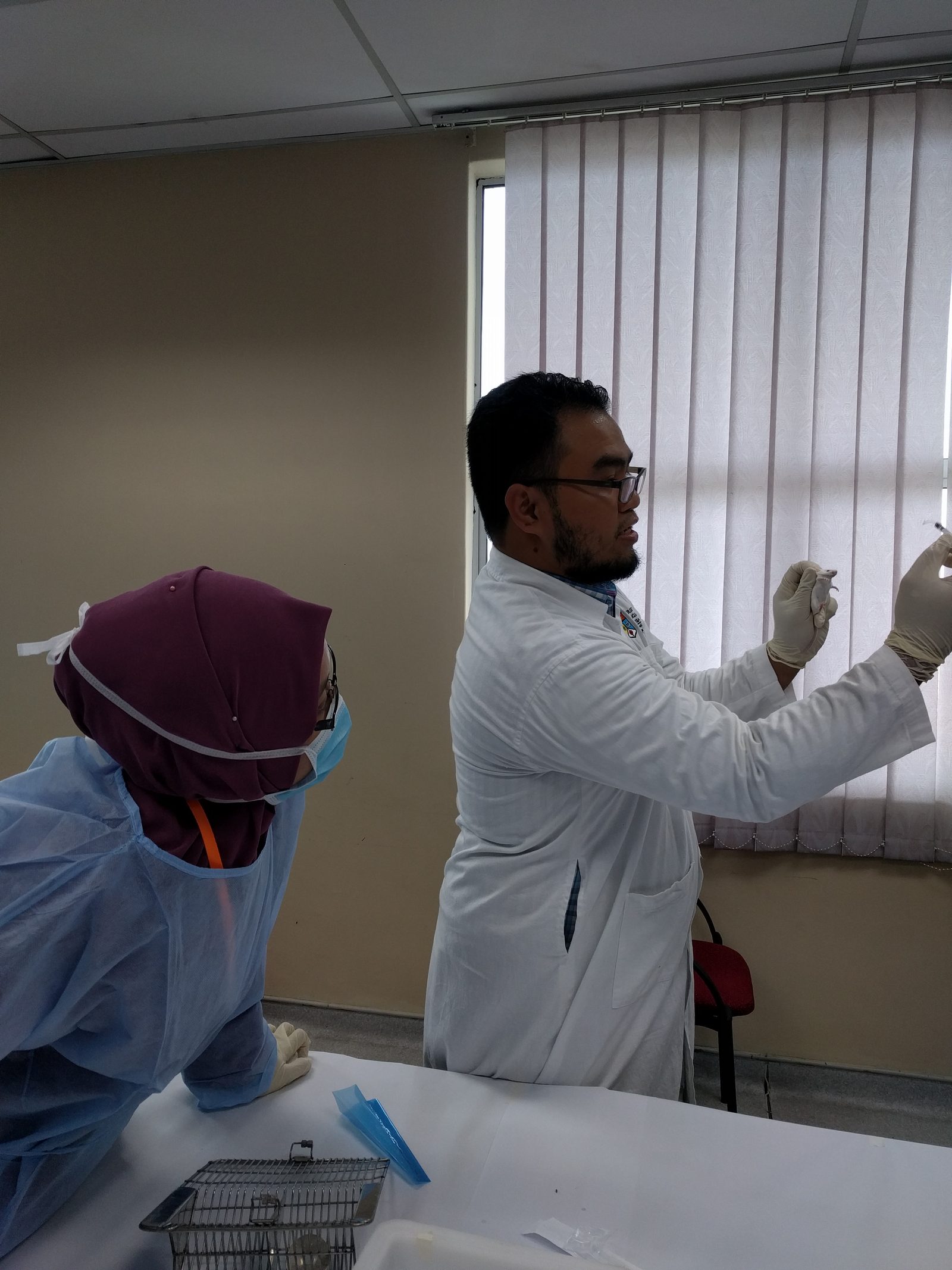 A man in a white lab coat holds up a syringe while a participant in a gown and mask watches
