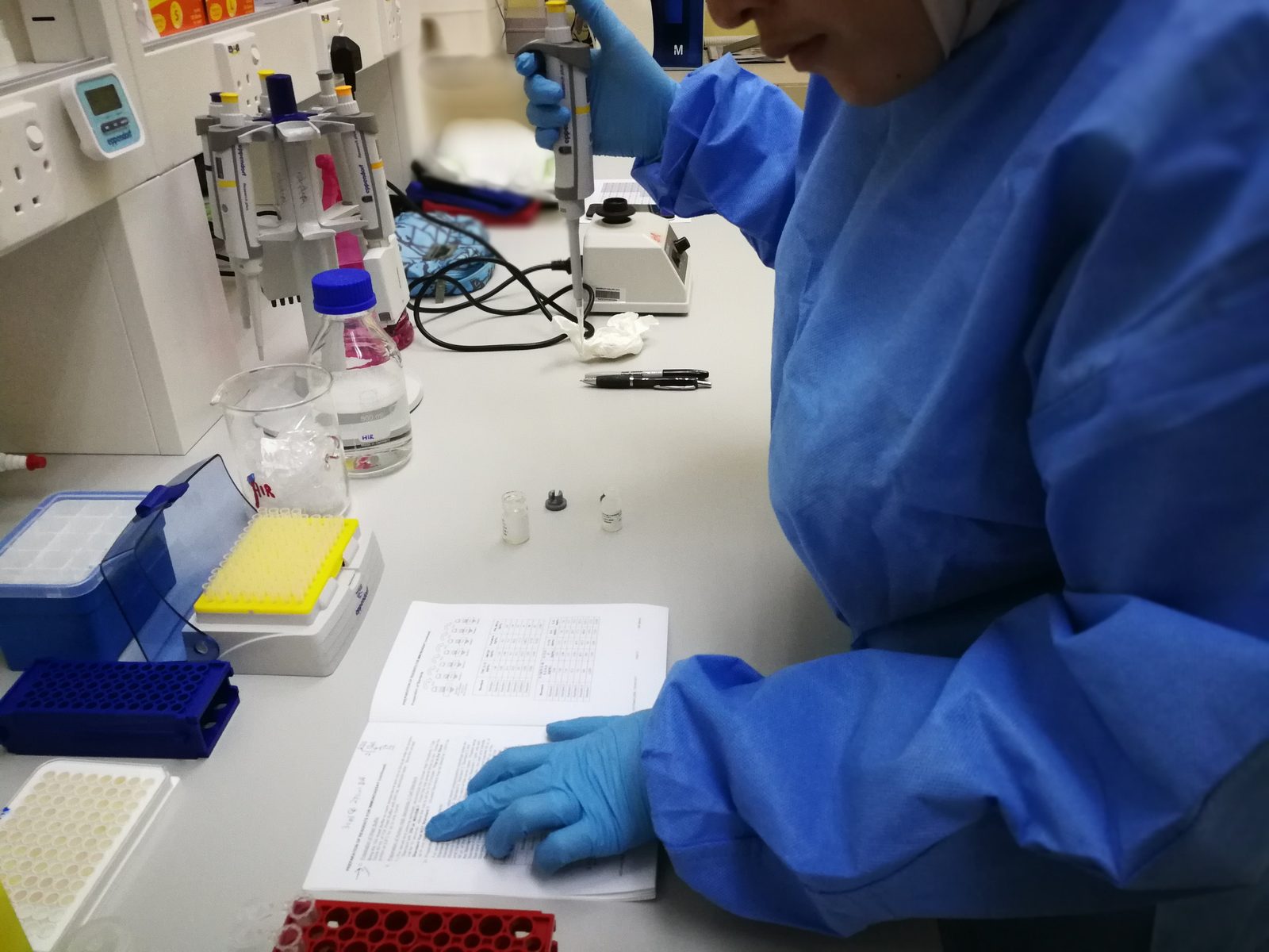 Gloved hands hold a pipette over a kit protocol at a lab bench, beside tube racks, reagent vials and a well plate.