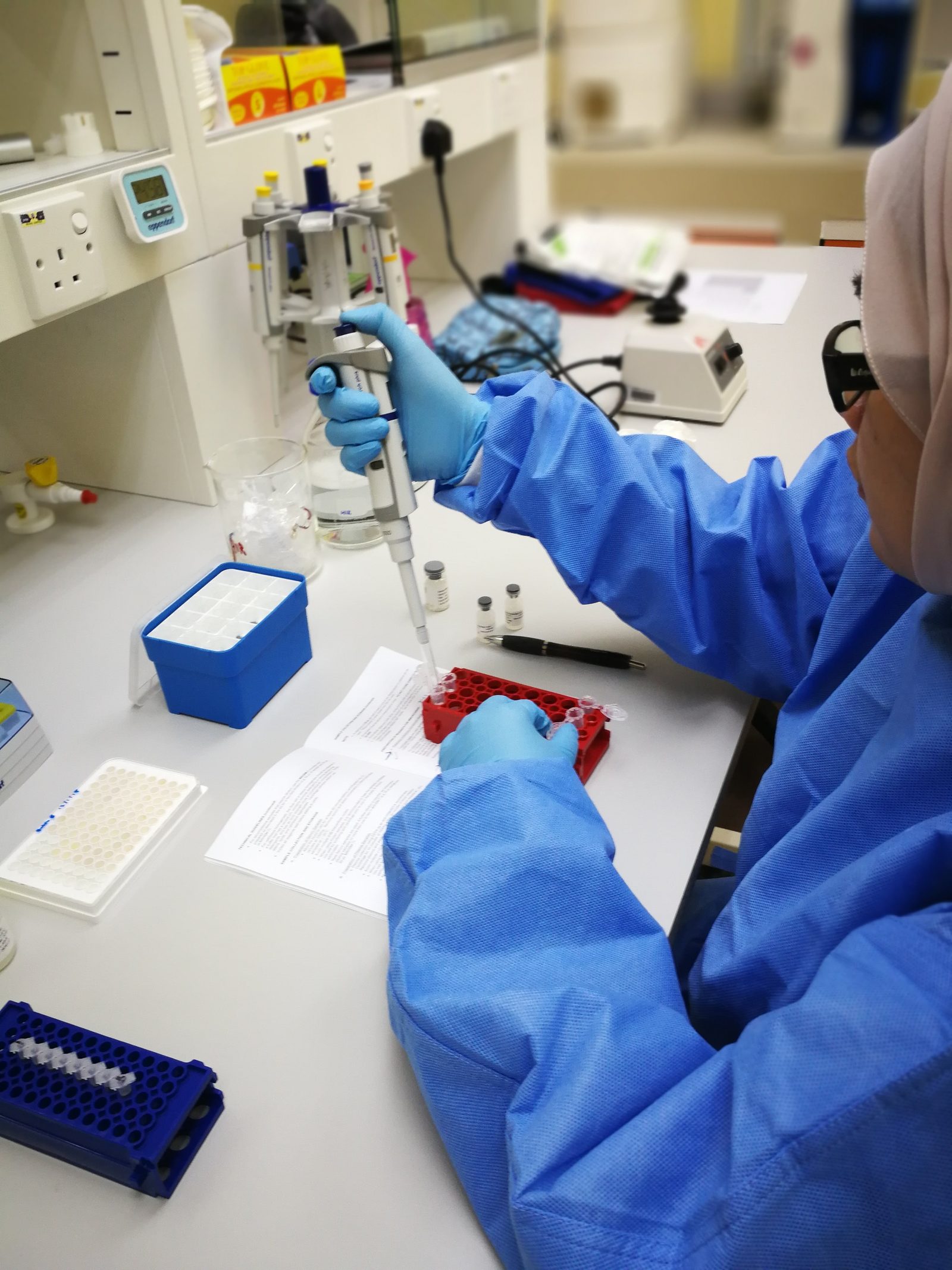 A researcher in a blue lab gown and gloves pipettes into a tube in a red rack, with the kit protocol open on the bench.