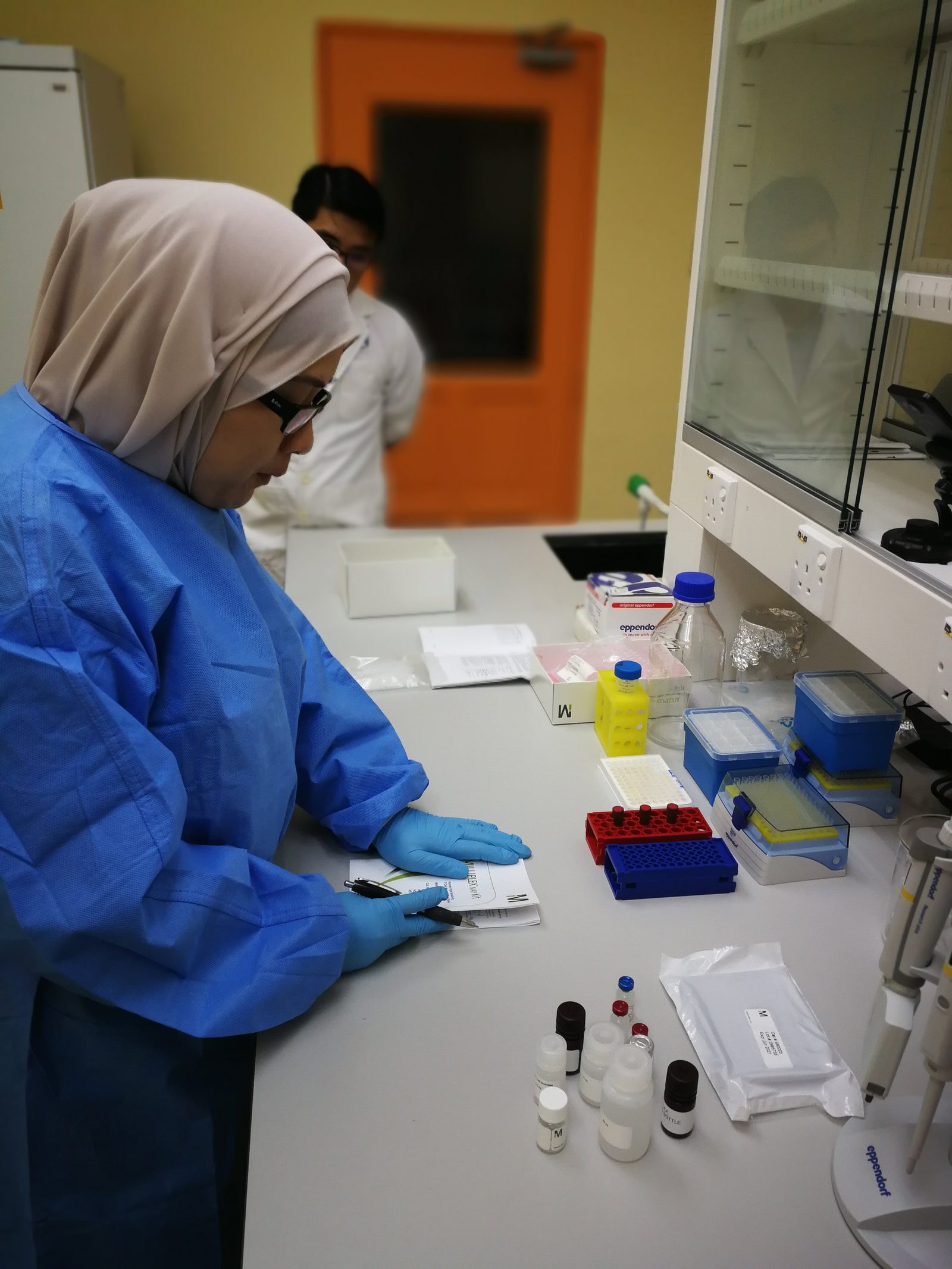 A researcher in a blue lab gown reads a kit insert at the bench while a man in a white coat looks on behind her.