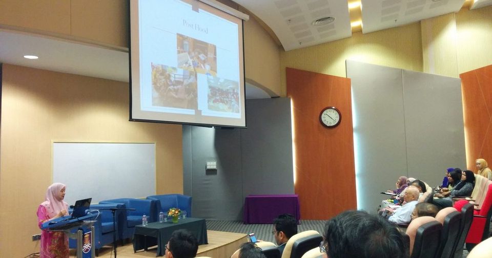 A speaker at the lectern presents a slide titled Post Flood to an audience in a university auditorium