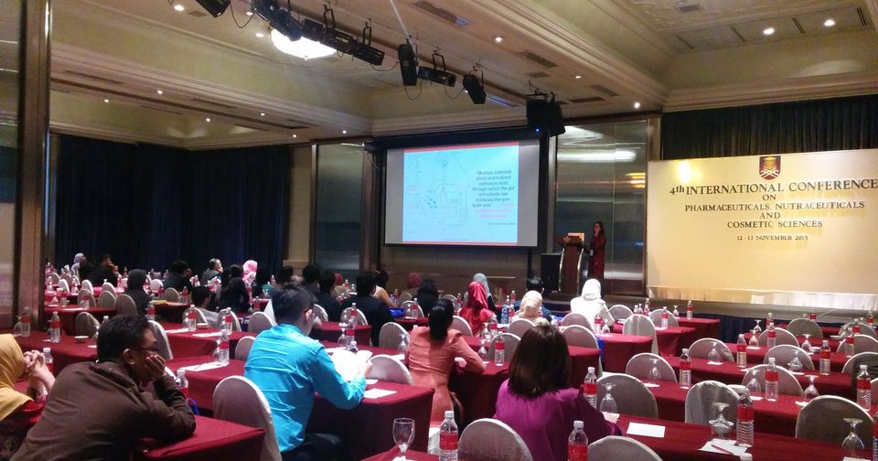 Delegates seated at rows of tables in a hotel ballroom, facing a screen and the conference banner