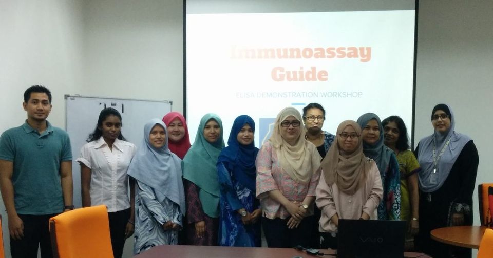 Workshop participants stand together in a meeting room in front of a slide titled Immunoassay Guide