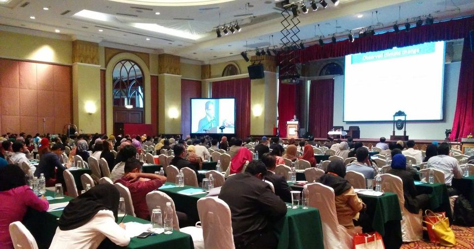 Delegates seated at long tables in a hotel hall, facing a speaker at the lectern and two large screens